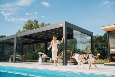 Woman walking a dog by a modern PERGOLUX pool house with glass walls.