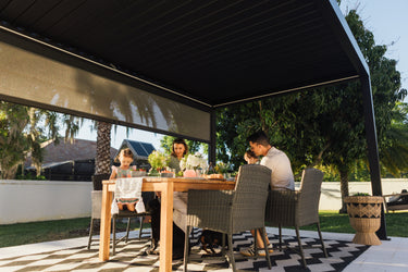 Family sitting at an outdoor dining table under a large PERGOLUX pergola on a sunny day.