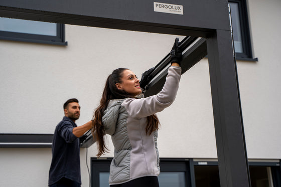 Woman installing pergola with a man assisting her, set against a building exterior.