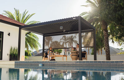 A modern outdoor patio cabana with a slatted roof and side screens, featuring two people sitting and dining under it by a swimming pool with palm trees and a house in the background.