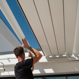 Person installing white louvers on a pergola with blue sky in the background