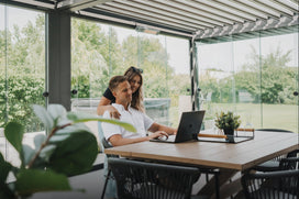 Two people working on a laptop under a modern pergola with a view of greenery outside.