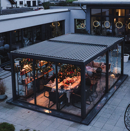 Outdoor dining area under a metal pergola with glass walls on a patio