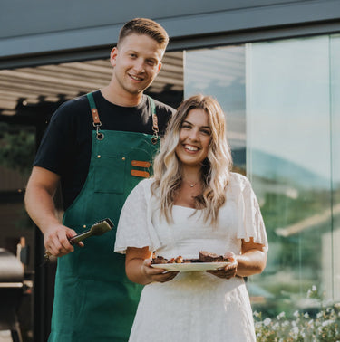 Man in a green apron and woman in a white dress holding food outdoors.